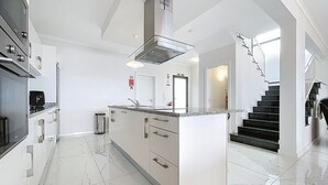 A modern kitchen with white cabinetry, a central island, stainless steel appliances, and a staircase leading to another level. - Blue Outlook Retreat by Atlantic Holiday (Santa Cruz)