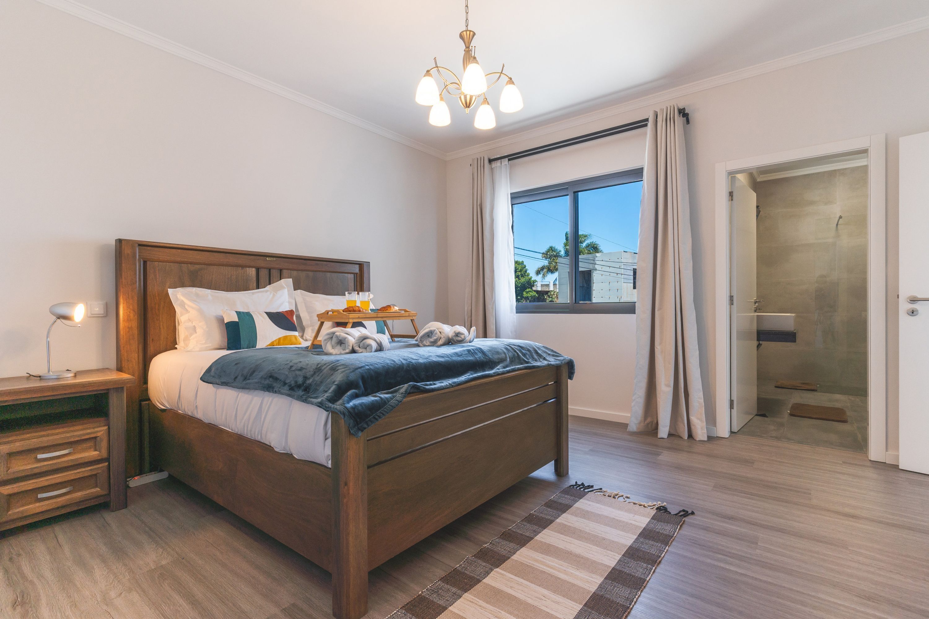 A modern bedroom with a wooden bed, bedside tables, a nightstand, a window with curtains, and a shower area.