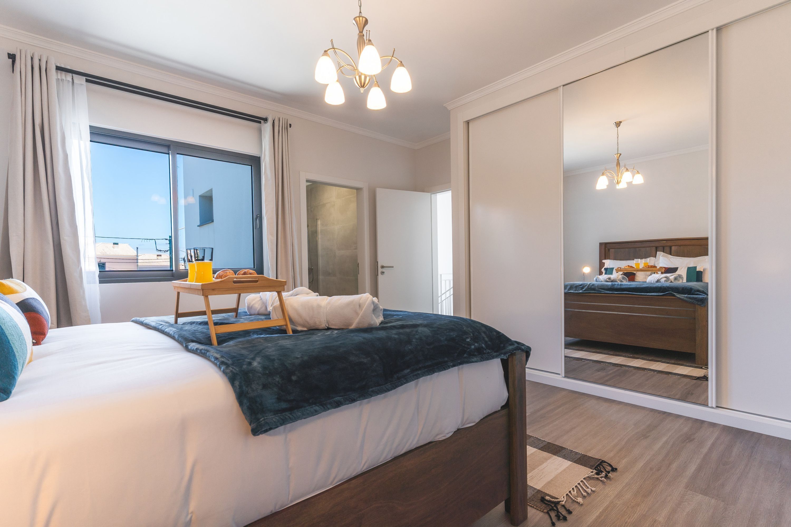A modern hotel room with a large bed, a bedside table with a lamp, a mirror, and a view of the city through a large window.