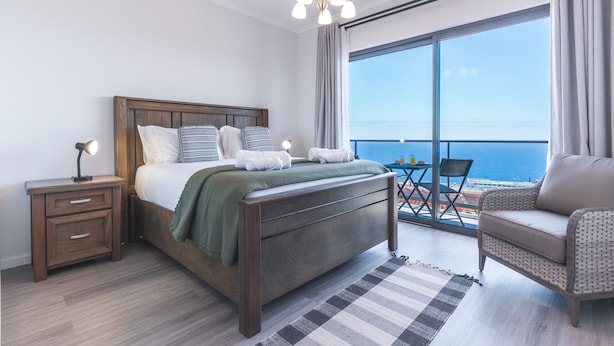 A bedroom with a large bed, a nightstand, a chair, and a view of the ocean through a large window. - Blue Outlook View by Atlantic Holiday (Santa Cruz)