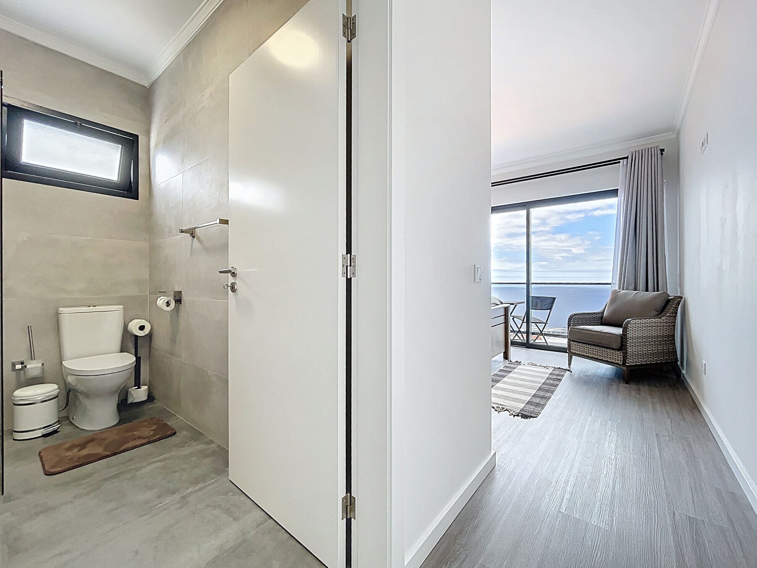Une salle de bain moderne équipée de toilettes, d’un bidet et d’un porte-serviettes. Un couloir mène à un balcon où se trouvent une chaise et une vue sur la mer.