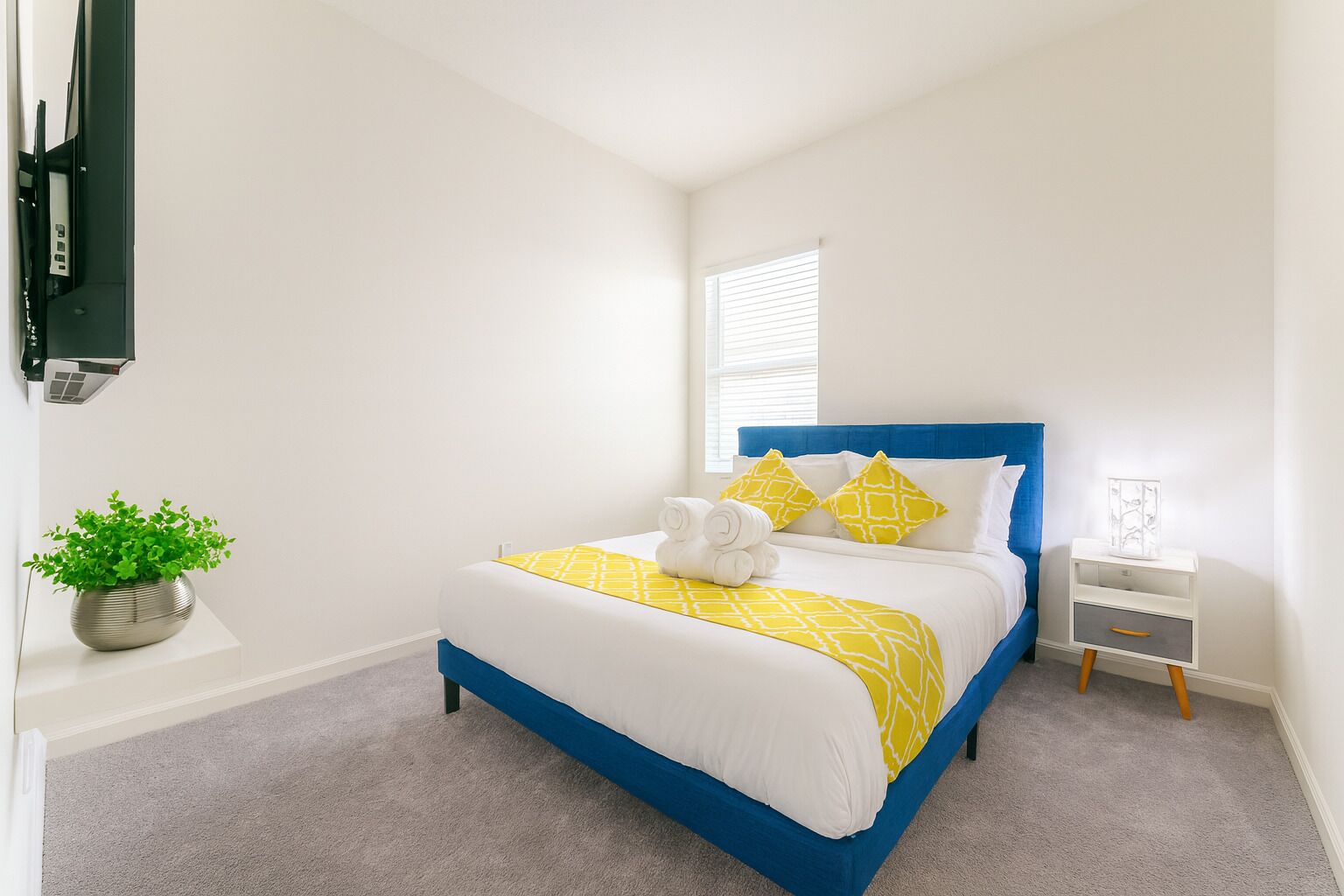 A neatly arranged bedroom with a bed, a bedside table, a nightstand, a potted plant, and a window with blinds.
