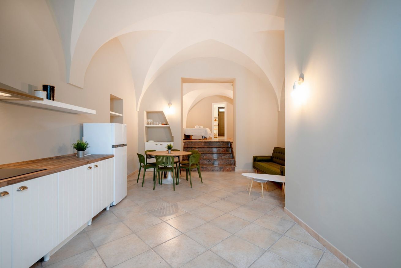 A modern kitchen with white cabinetry, a dining area with green chairs, and a staircase leading to a bedroom.