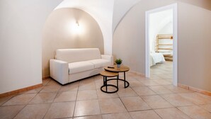 Una sala de estar moderna con un sofá blanco, una mesita redonda y un acceso a un dormitorio con cama y mesita de noche.