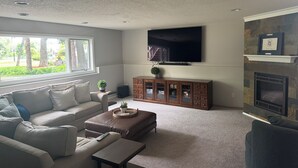 A living room with a sofa, ottoman, and coffee table, a flat-screen TV mounted on the wall, and a fireplace with a mantel. -  3M Golf  Immaculate Spacious 5-bedroom house in  Blaine. (Blaine)