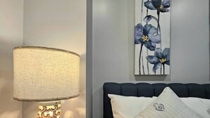 Una cama bien hecha con un cabecero oscuro y almohadas estampadas, una lámpara de noche con un pie decorativo, y una obra de arte floral en la pared.