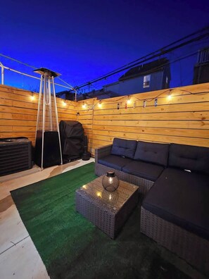 Un patio con un muro de madera, una parrilla negra, un sofá y una mesa de vidrio.