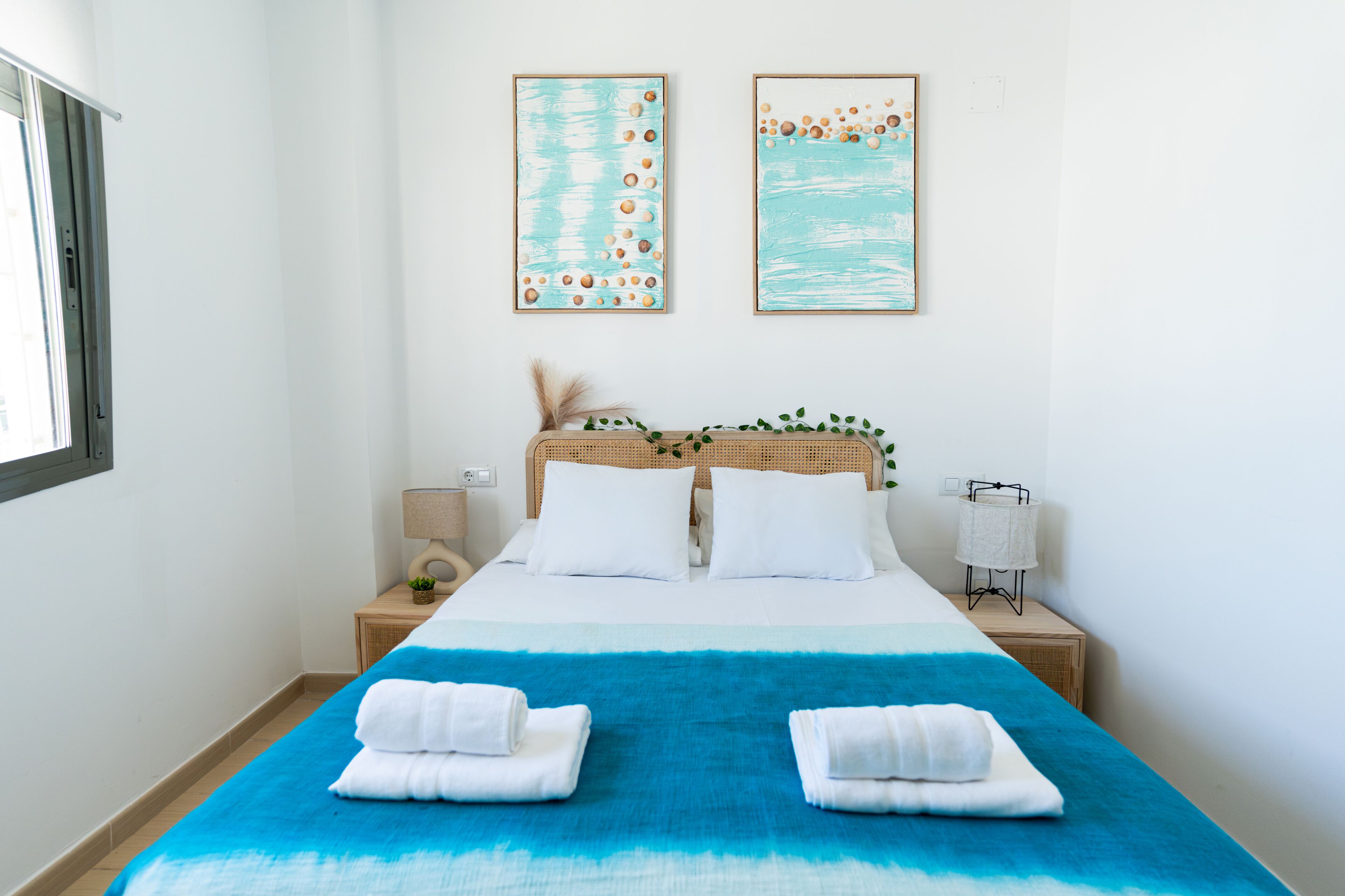 Quarto de hotel com uma cama grande, duas obras de arte na parede, uma mesa de cabeceira com abajur e uma janela.