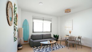 Una sala de estar moderna con un sofá gris, una mesa redonda de centro, una mesa de comedor con sillas, una tabla de surf y un gran ventanal con persianas.