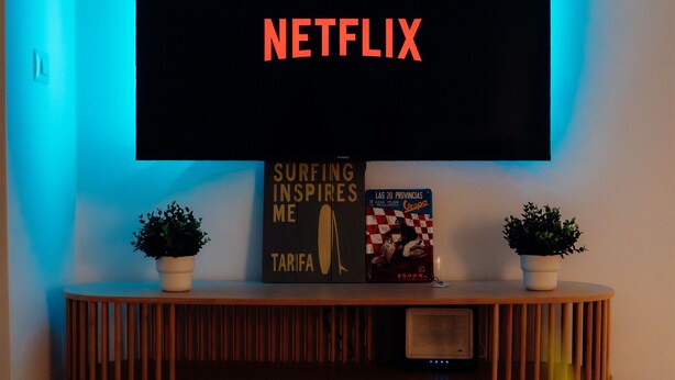 Una sala de estar moderna con un televisor de pantalla plana grande montado en la pared, una consola de madera con dos plantas en macetas, y un libro titulado "Surfing Inspires Me" de Tarifa.