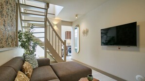 Een moderne woonkamer met een hoekbank, een kleine ronde tafel, een aan de muur bevestigde televisie en een trap naar een andere kamer.