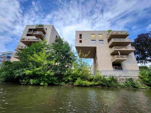 Un edificio moderno con varios balcones, un río al frente y abundante vegetación alrededor.