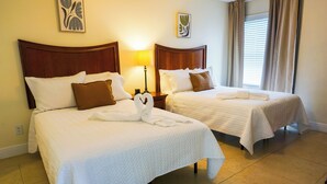 Two double beds with white linens and brown pillows, a nightstand with a lamp, and artwork on the walls.