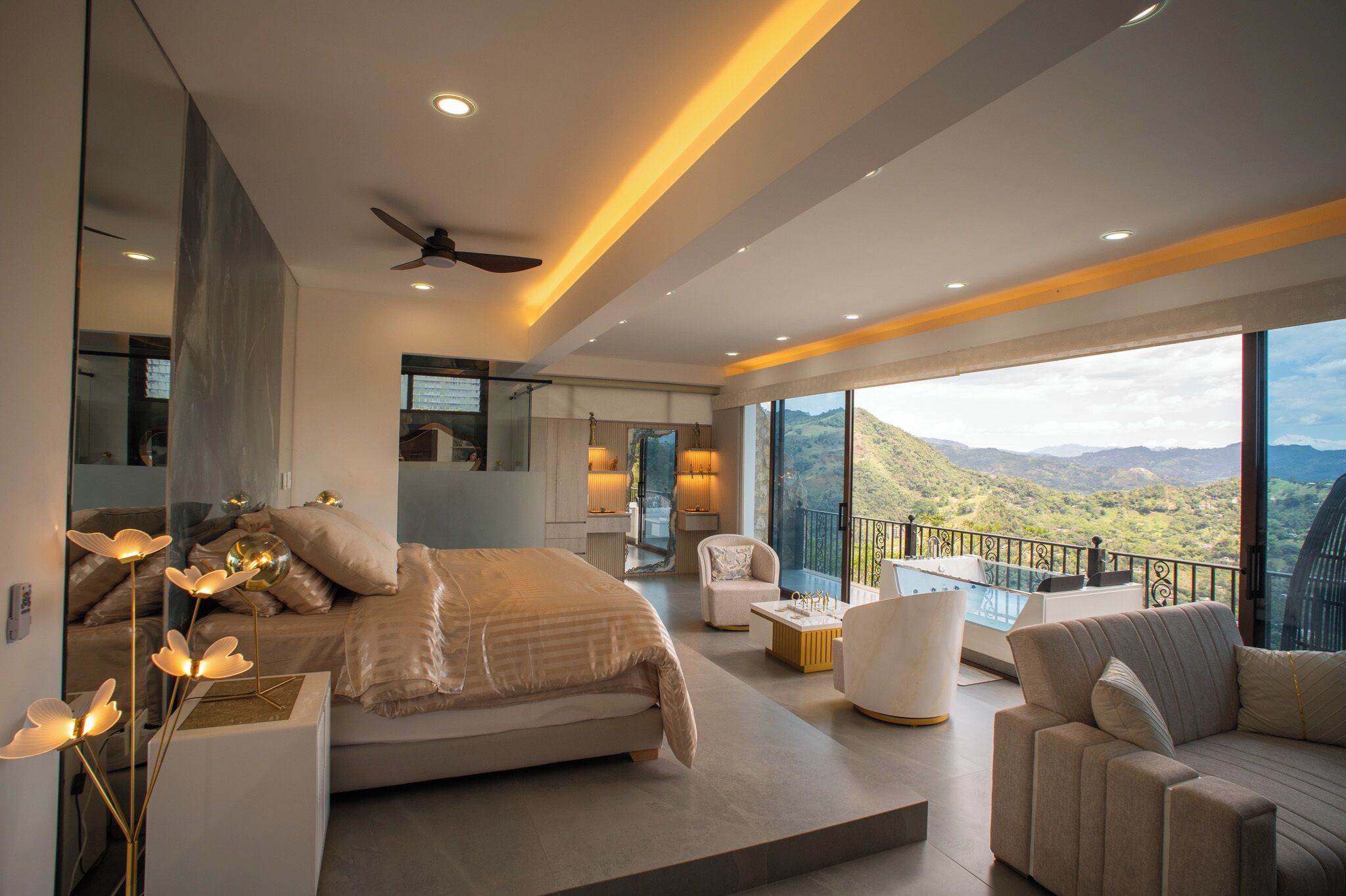 Un dormitorio moderno con una cama grande, una zona de estar con sofá y sillones, un balcón con vistas a las montañas y un baño con bañera.