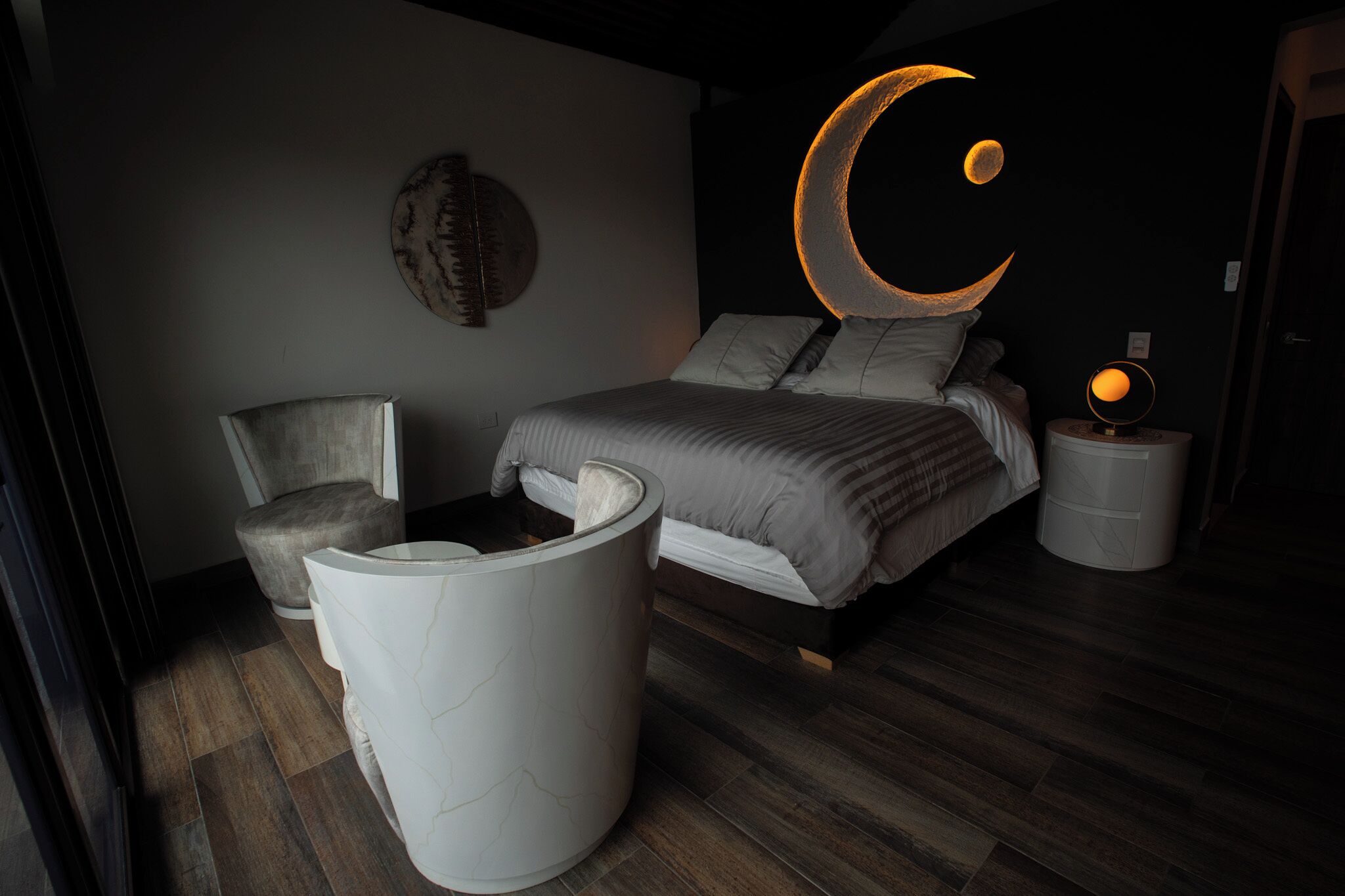 A modern bedroom with a bed, a chair, a bedside table, and a wall-mounted light fixture.