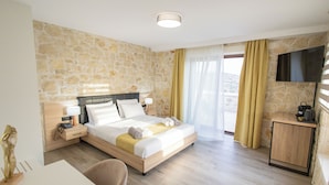 Habitación de hotel con pared de piedra, cama con ropa blanca, mesita de noche de madera con lámpara, televisor montado en la pared y vista al exterior a través de una ventana con cortinas.