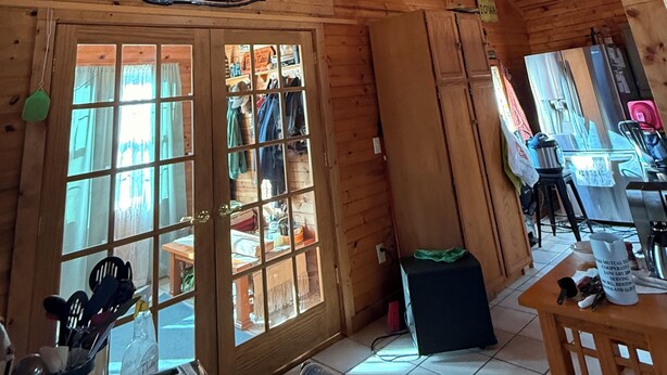 A wooden interior with a high ceiling, a glass door leading outside, and a visible kitchen area with a refrigerator and table.
