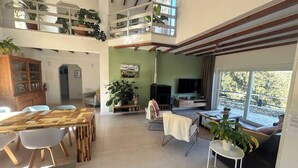 Un vaste espace de vie doté d’un haut plafond, de poutres apparentes, d’une table à manger avec des chaises, d’un canapé, d’un téléviseur et de nombreuses plantes en pot.