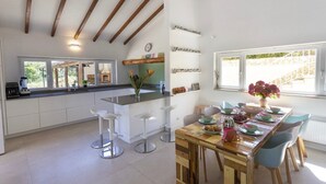 Une cuisine moderne avec des armoires blanches, un îlot central en bois et des tabourets de bar. On y trouve également un coin repas avec une table et des chaises en bois, ainsi qu’une fenêtre donnant sur l’extérieur.