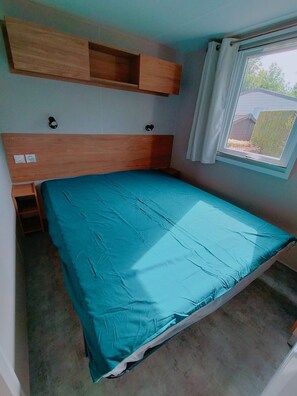 Una habitación pequeña con cama individual, cabecera de madera, ventana con cortinas y vista al exterior.