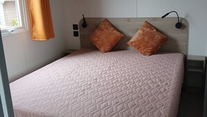 Une chambre à coucher comprenant un lit, deux oreillers, une table de chevet, une étagère avec des objets décoratifs et une fenêtre avec des rideaux.