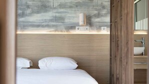 A hotel room with a wooden headboard, a bed with white linens, a wall-mounted light, and a visible bathroom with a sink and mirror.