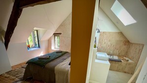 Ein Schlafzimmer mit schrägen Wänden, einem Bett, einem Fenster und Blick ins Freie. Es ist auch ein Badezimmer mit einer Badewanne, einem Waschbecken und einer Dusche zu sehen.