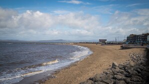 A beach with a rocky shoreline, a building with a flat roof, and a distant mountain range. - Altham Place-fully Refurbished 2-bath Bungalow (Morecambe)
