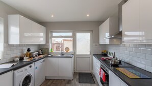 A modern kitchen with white cabinets, a built-in washing machine, a sink, and a window with a view of the outside. - Altham Place-fully Refurbished 2-bath Bungalow (Morecambe)