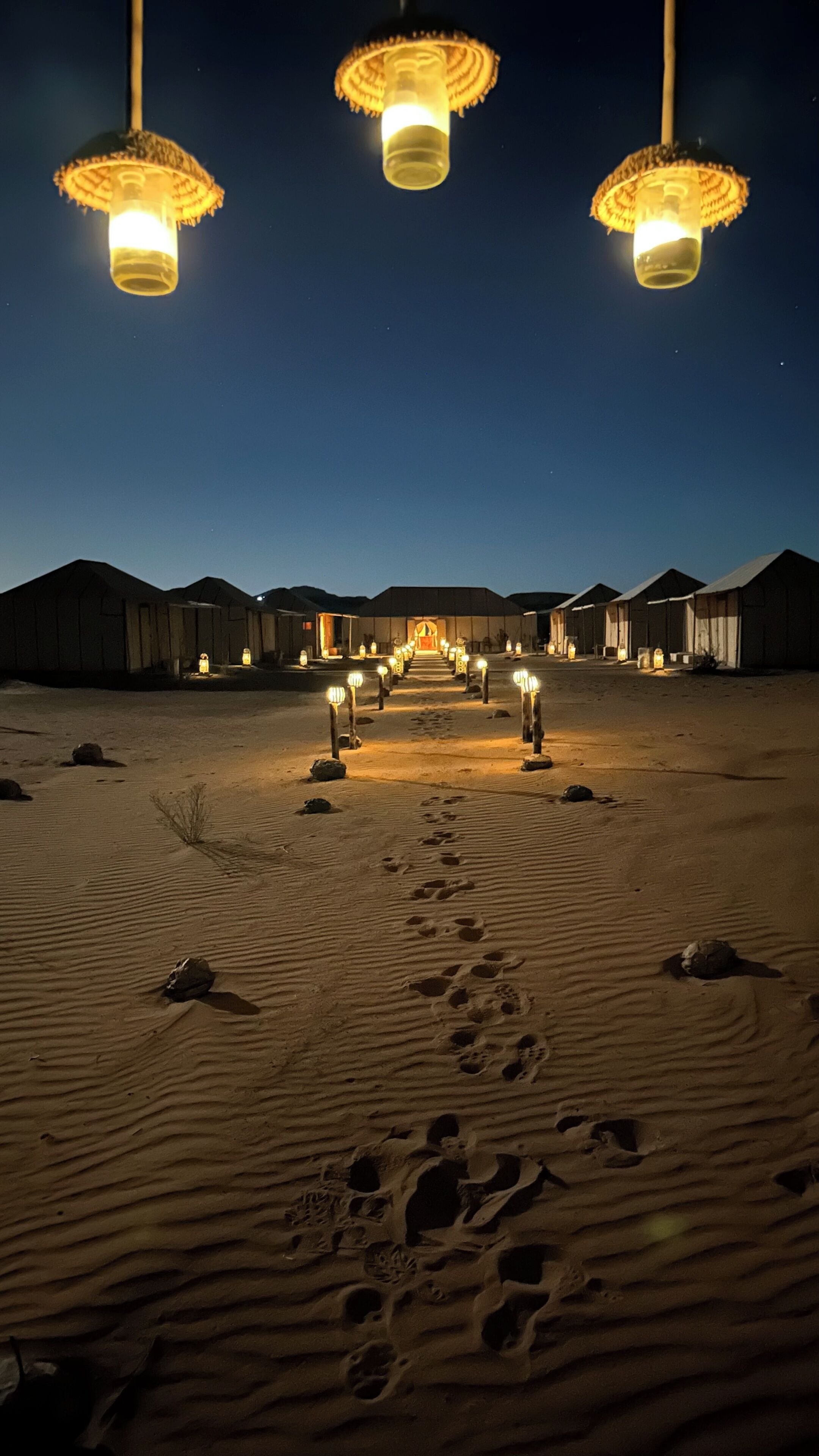 A desert hotel with a central pathway leading to a building, surrounded by tents, under a night sky.