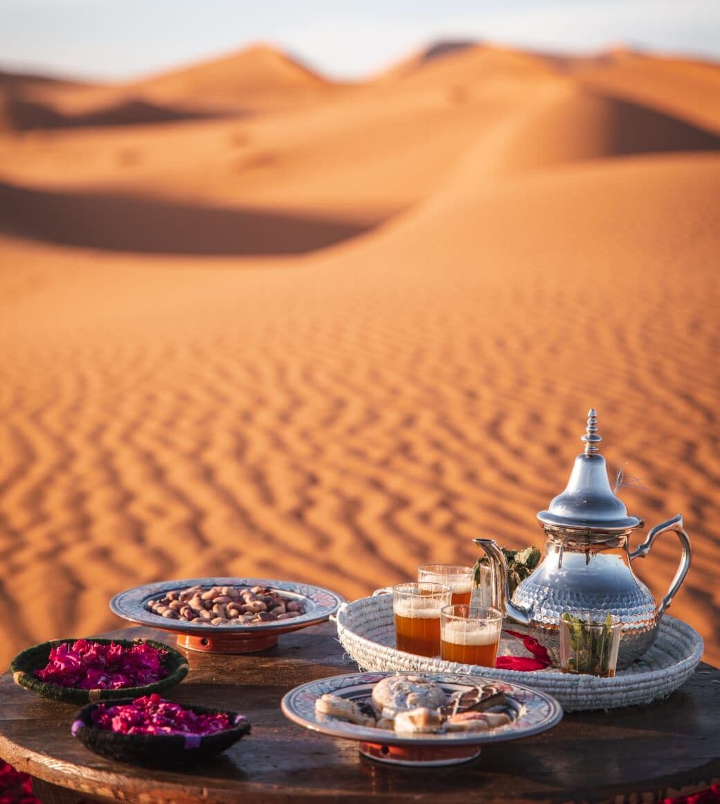 A desert scene with a table set with traditional Moroccan tea and snacks, a metal teapot, and a woven tray.