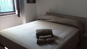 Un lit bien fait, recouvert d’une couvre-lit beige et agrémenté d’oreillers, une fenêtre ornée de barreaux en fer décoratifs et une petite table de chevet avec une lampe.