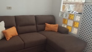 Un canapé d’angle marron avec deux coussins, un blanc et un orange, dans une pièce dotée d’une fenêtre et d’un rideau à motifs.