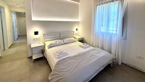 A bedroom with a bed, bedside tables, a ceiling light, a window with curtains, and a wooden ceiling beam.