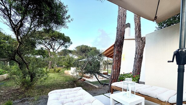 An outdoor patio with white cushioned seating, a small table, and a hammock. Trees and a building are visible in the background.