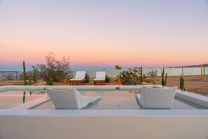 Área da piscina com espreguiçadeiras e vista para o mar ao pôr do sol.