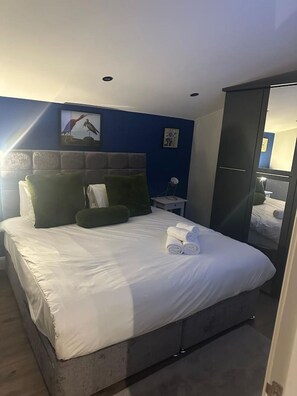 A neatly made bed with white linens and green pillows, a bedside table with a lamp, and a wall-mounted picture of a bird.
