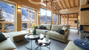 Un salón amplio con un sofá grande, una mesa de centro y una chimenea. Amplias ventanas ofrecen vistas a un paisaje montañoso nevado.