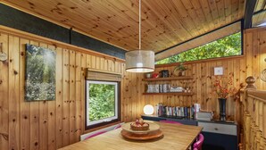 Een houten interieur met een eettafel, roze stoelen, een hanglamp, een raam en planken met boeken en decoratieve objecten.