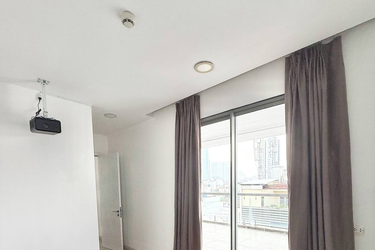 A hotel room with a large bed, a projector mounted on the ceiling, and a view of the city through the window.