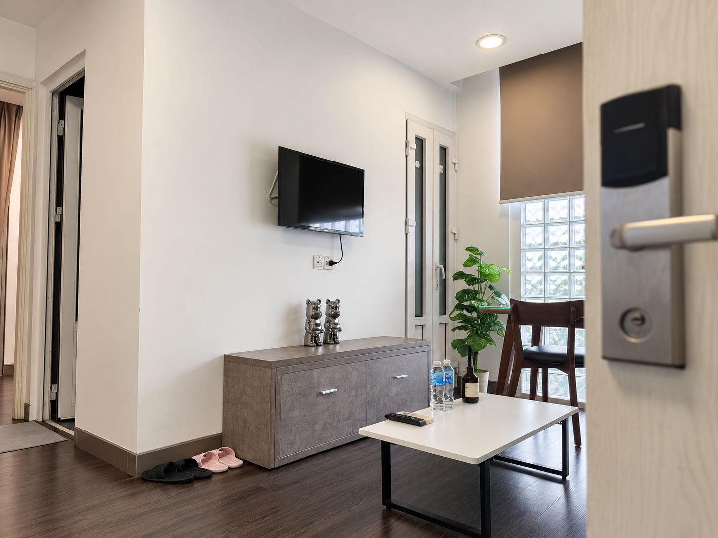 A modern room with a flat-screen TV mounted on the wall, a grey cabinet, a small table with a remote control, a potted plant, and a wooden chair.