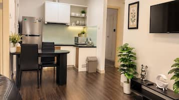 Een moderne woonkamer met een leren bank, een eethoek met een zwarte tafel en stoelen, een keuken met witte kasten en roestvrijstalen apparatuur, een aan de muur bevestigde televisie, en een potplant.