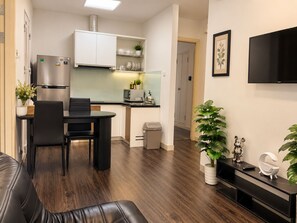 Uma sala de estar moderna com sofá de couro, área de jantar com mesa e cadeiras pretas, cozinha com armários brancos e eletrodomésticos de aço inoxidável, televisão na parede e uma planta em vaso.