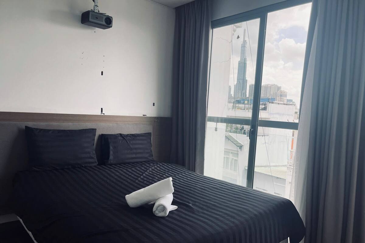A hotel room with a large bed, a projector mounted on the wall, and a view of a cityscape through a large window.
