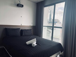 A hotel room with a large bed, a projector mounted on the wall, and a view of a cityscape through a large window. - Uhouse - MELIA Thao Dien Hotel & Apartment (Ho Chi Minh City)