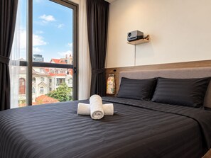 Habitación de hotel con una cama grande, un proyector en la pared y vistas a edificios al exterior.