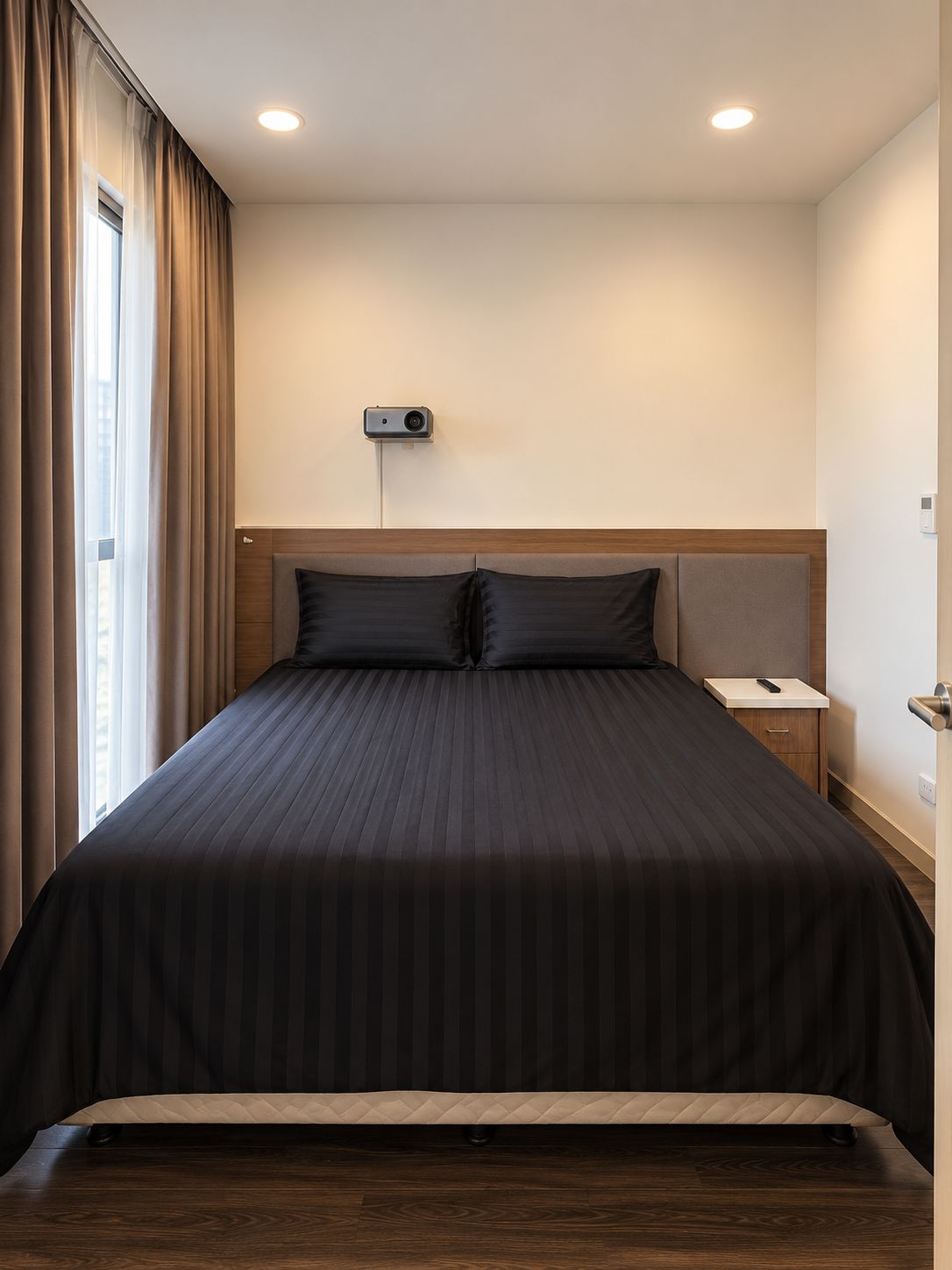 Habitación de hotel con una cama grande, una mesita de noche, un proyector en la pared y una ventana con cortinas.