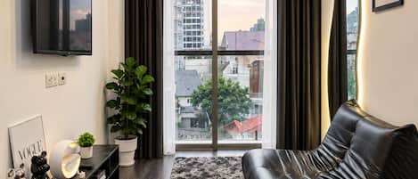 A modern living room with a black leather sofa, a flat-screen TV mounted on the wall, a potted plant, and a view of the cityscape through large windows.