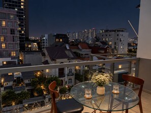 Un balcón con una mesa y sillas de vidrio, con vistas a un paisaje urbano nocturno.