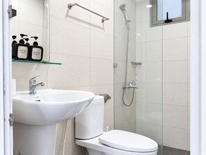 Une salle de bain moderne avec un lavabo, des toilettes et une douche, le tout de couleur blanche. On y trouve une étagère avec des articles de toilette et une paroi de douche en verre.
